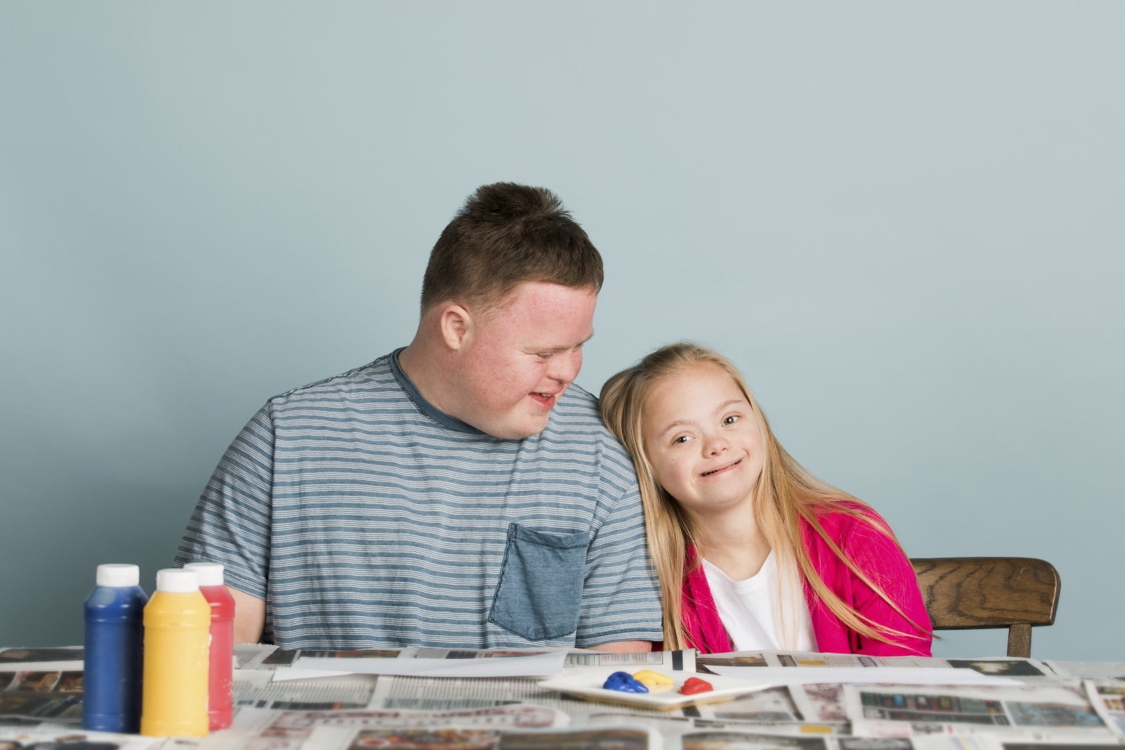 Two children with Down syndrome are seated at a table covered with newspapers, paint bottles, and art supplies. The boy is wearing a striped shirt, and the girl is wearing a pink cardigan—truly reflecting the joy that empowerment care services aim to bring into their lives.