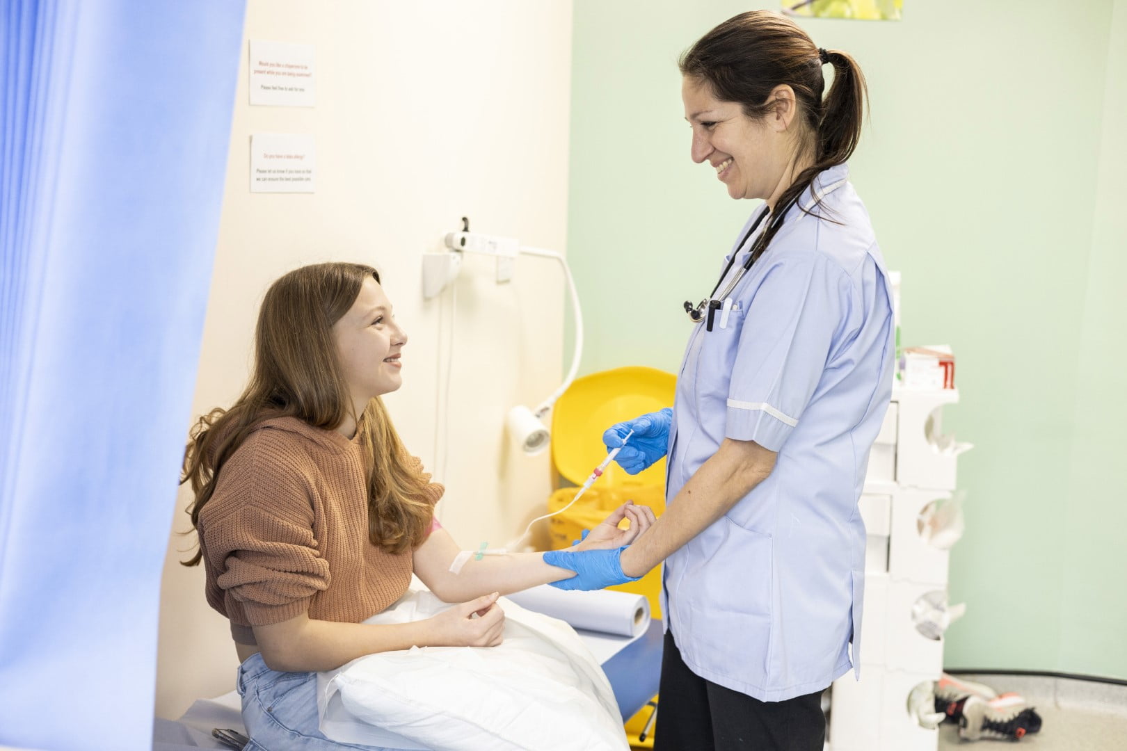 A nurse wearing blue gloves and a stethoscope administers an IV to a smiling female patient seated on a hospital bed, highlighting the compassionate support of Community Nursing Care.