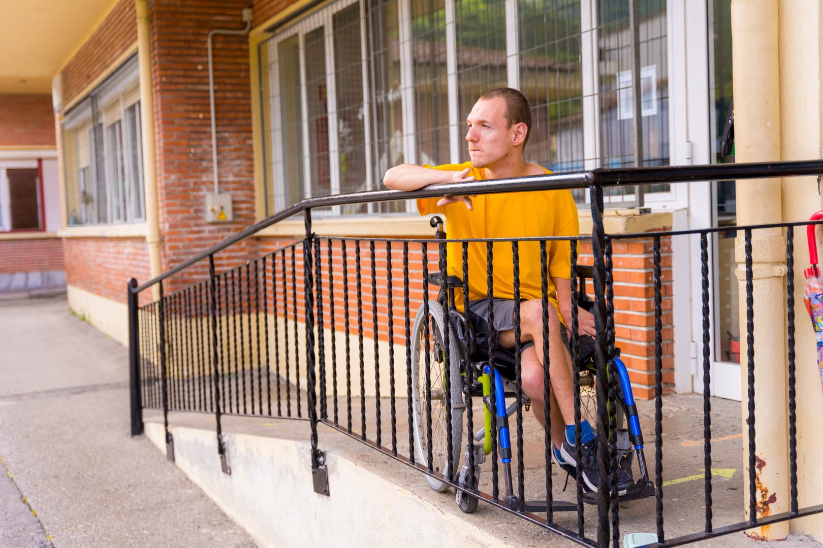 A person in a yellow shirt sits in a wheelchair at the top of a ramp outside a brick building, part of the Specialist Disability Accommodation (SDA) complex. They are looking towards the distance.