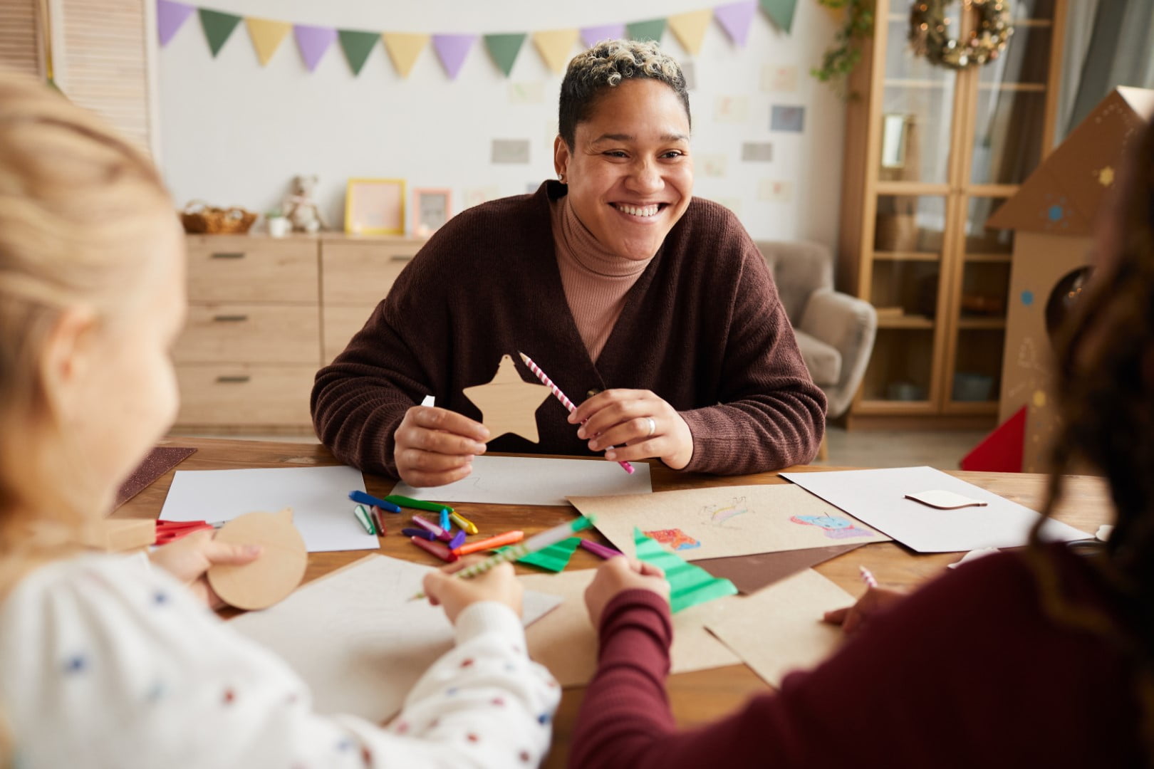 A person smiles while crafting with two children at a table, surrounded by various papers and colored pencils, enjoying the creative moment provided by respite services.