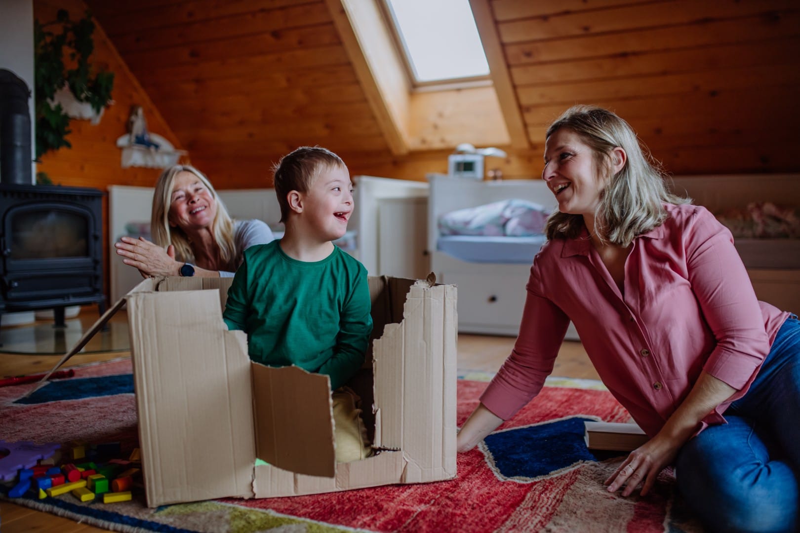 A young boy sits in a cardboard box on a rug, smiling at two women nearby in a cozy, wood-paneled room with toys scattered on the floor—an uplifting scene highlighting joyful moments supported by Select NDIS Services.