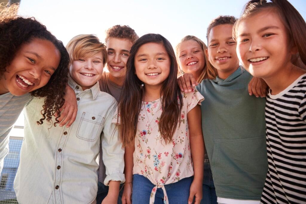 A group of seven smiling children in Specialised Substitute Residential Care (SSRC) stand close together outdoors, posing for a photo on a sunny day.