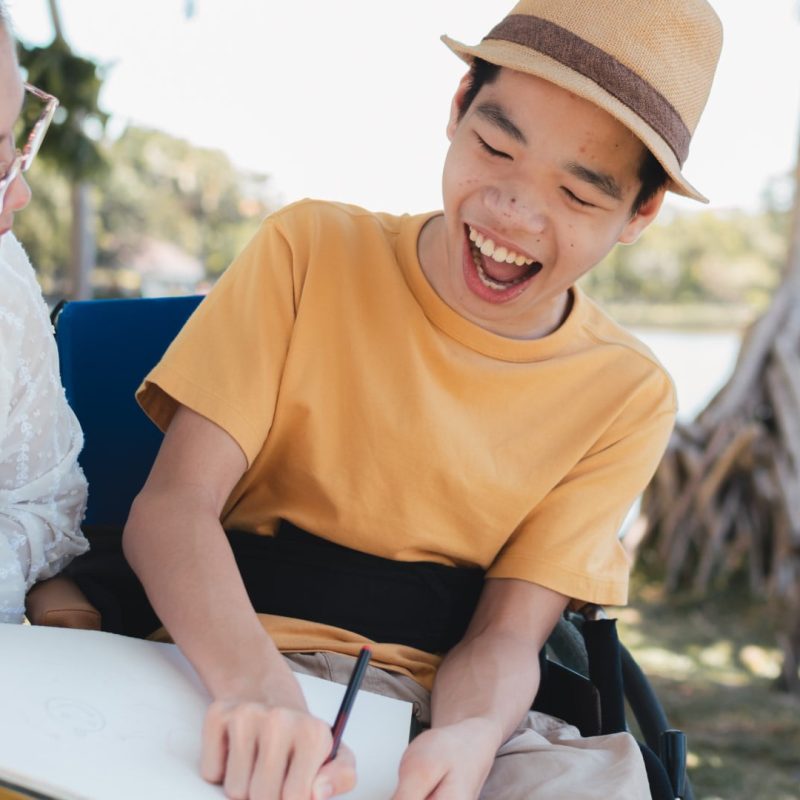A young person in a yellow shirt and hat is laughing while drawing in a sketchbook, sitting next to another individual who is also engaged in the activity, illustrating the joy and empowerment care services can bring.