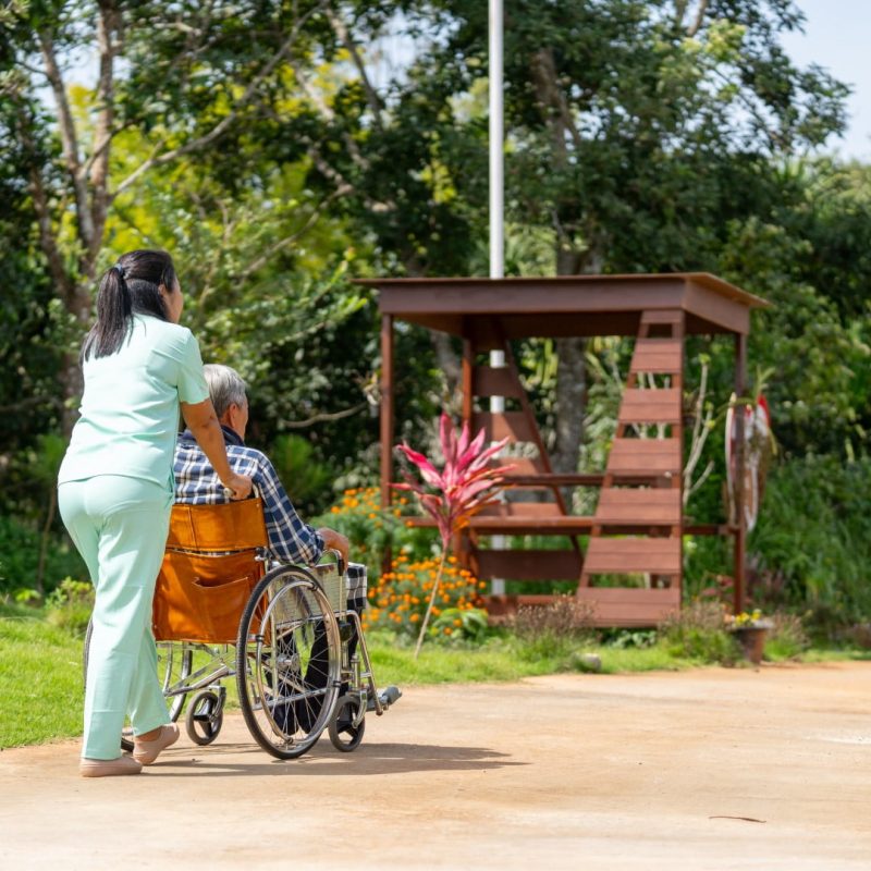 A caregiver in a light green uniform is pushing a person in a wheelchair along a pathway in a lush garden with a wooden gazebo, highlighting the quality of life available through Specialist Disability Accommodation (SDA).