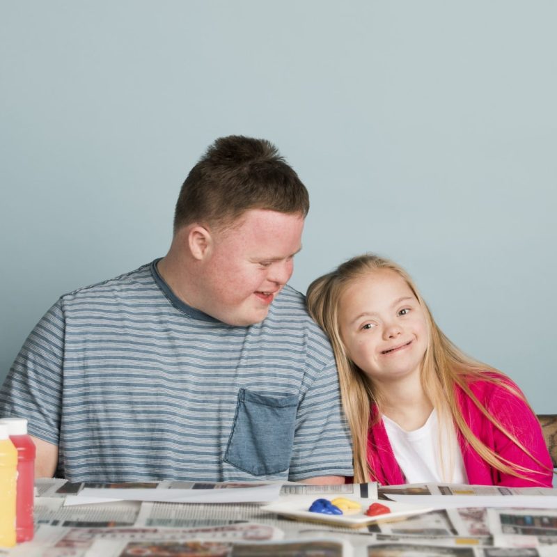 Two children with Down syndrome are seated at a table covered with newspapers, paint bottles, and art supplies. The boy is wearing a striped shirt, and the girl is wearing a pink cardigan—truly reflecting the joy that empowerment care services aim to bring into their lives.