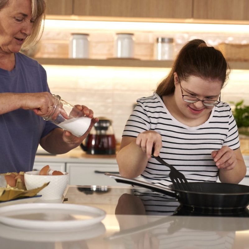 Two women are cooking in a modern kitchen. One is cracking eggs while the other, who benefits from Supported Independent Living (SIL), is stirring a pan on the stove. Various kitchen items are on the counter, showcasing their collaborative effort in a supported environment.