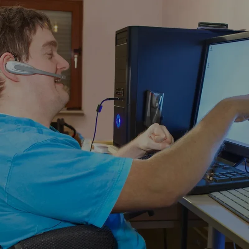 A person in a blue shirt using assistive technology, pointing at a computer screen with two monitors on a desk. They are wearing a headset and seated in a wheelchair.