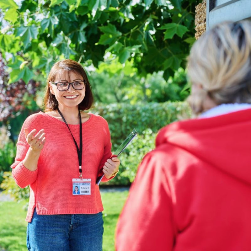 A woman wearing glasses, holding a clipboard, and sporting a name badge, engages in conversation with another woman dressed in red outdoors in a garden setting, discussing Community Nursing Care.