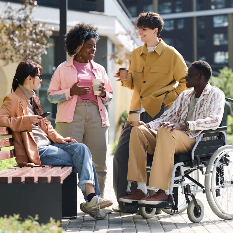 Four friends chat and smile together outdoors—one on a bench, one holding a drink, another with a coffee cup, and one using a wheelchair—showcasing ECS Community Innovation and empowered living amid lush greenery and modern buildings.