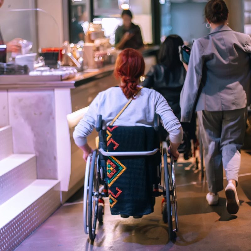 A person with red hair in a wheelchair, draped with a patterned blanket, moves through a modern, busy café with others—reflecting the spirit of ECS Community Innovation & Empowered Living.