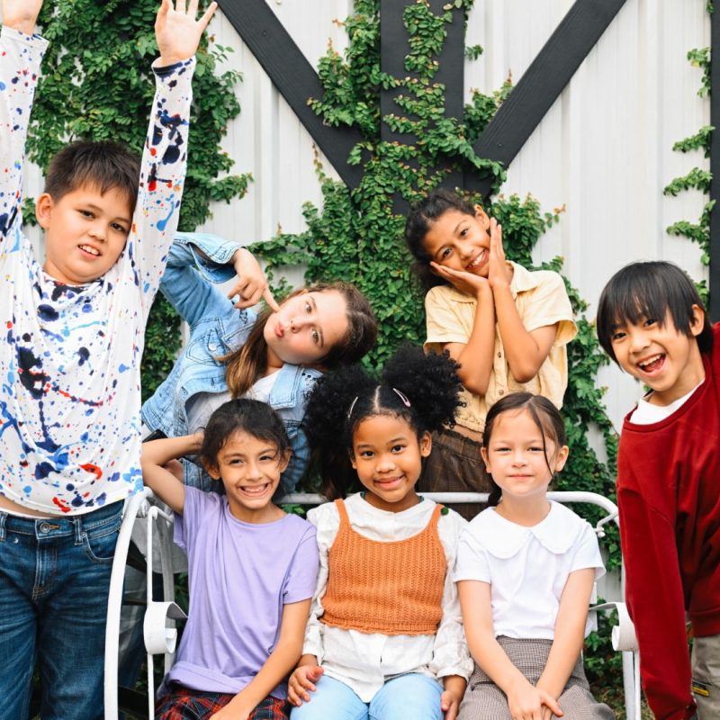 A group of seven children posing cheerfully in front of a wall with greenery, some standing and some sitting on a bench, with various joyful expressions and raised arms, embodying the spirit of empowerment care services.