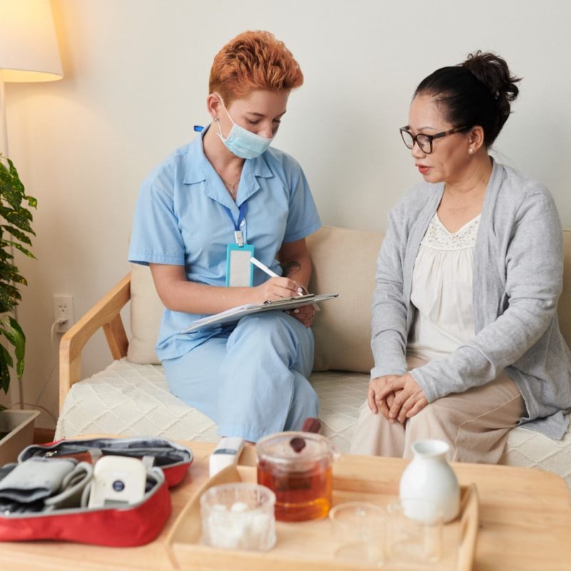 A healthcare worker in a blue uniform and mask speaks with a seated woman. They are on a couch in a room with a plant and a small table holding various medical supplies and a cup of tea, exemplifying the essence of community nursing care.
