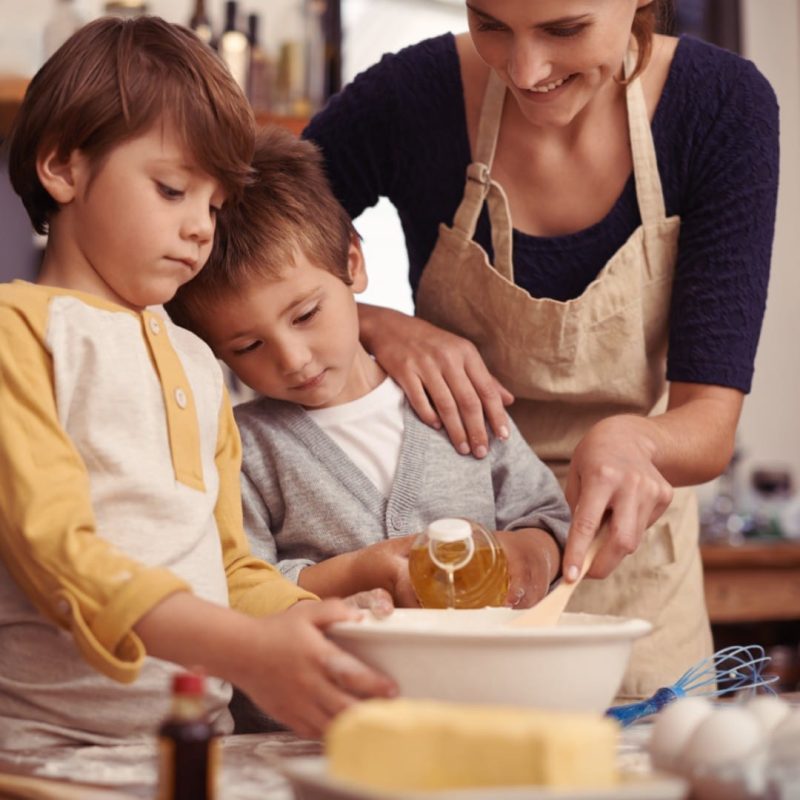 An adult and two children bake together, pouring oil into a mixing bowl in a kitchen with baking ingredients on the counter, enjoying quality time typical of Supported Independent Living (SIL).