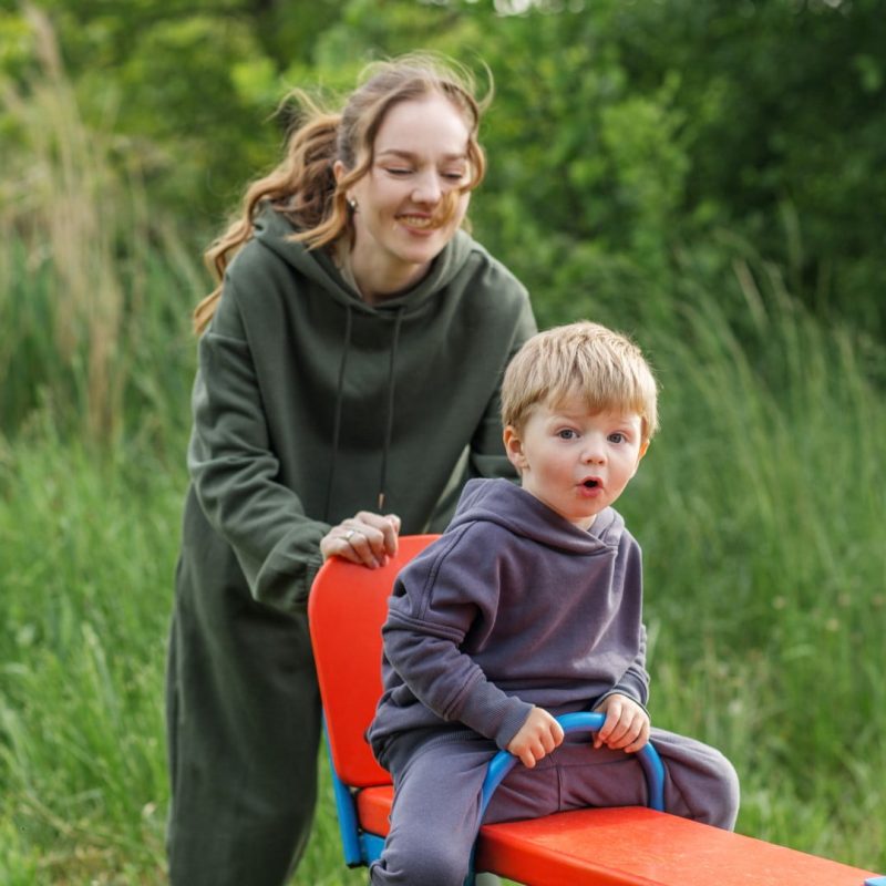 An adult and toddler in hoodies are on a seesaw in a grassy outdoor area, with the adult pushing the seesaw down.