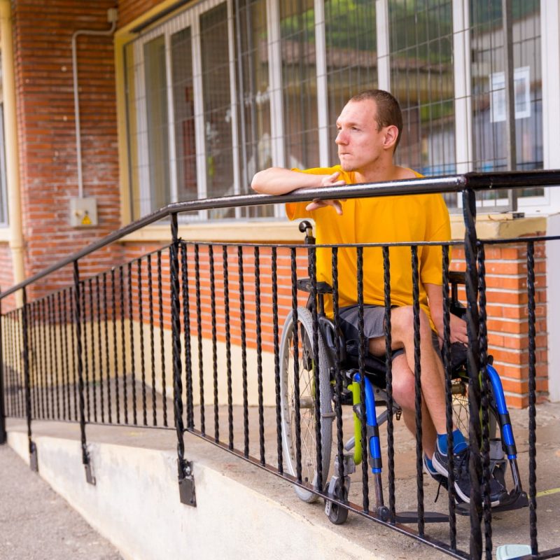 A person in a yellow shirt sits in a wheelchair at the top of a ramp outside a brick building, part of the Specialist Disability Accommodation (SDA) complex. They are looking towards the distance.