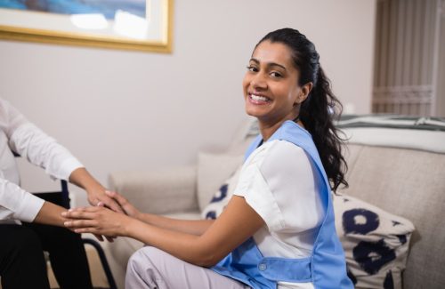 A woman in a blue apron sits on a couch, smiling at the camera, while holding the hands of another person sitting nearby, exemplifying the warmth and connection central to Community Nursing Care.
