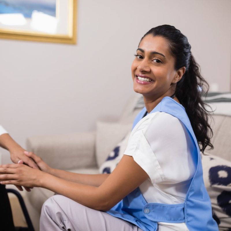 A woman in a blue apron sits on a couch, smiling at the camera, while holding the hands of another person sitting nearby, exemplifying the warmth and connection central to Community Nursing Care.