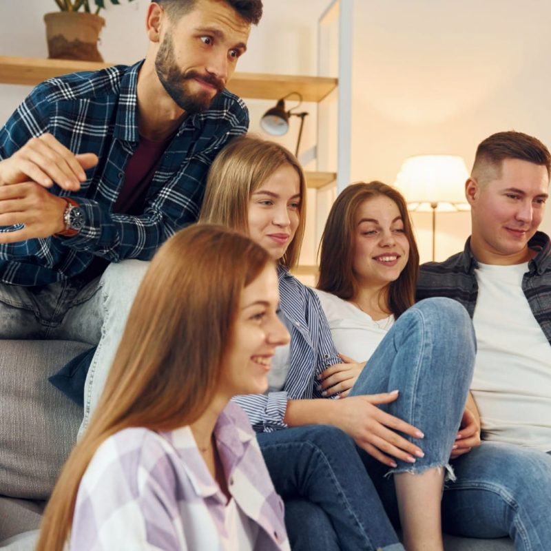 Five young adults sit close together on a couch in a living room, appearing relaxed and focused, possibly watching television in their Specialised Substitute Residential Care (SSRC) home.