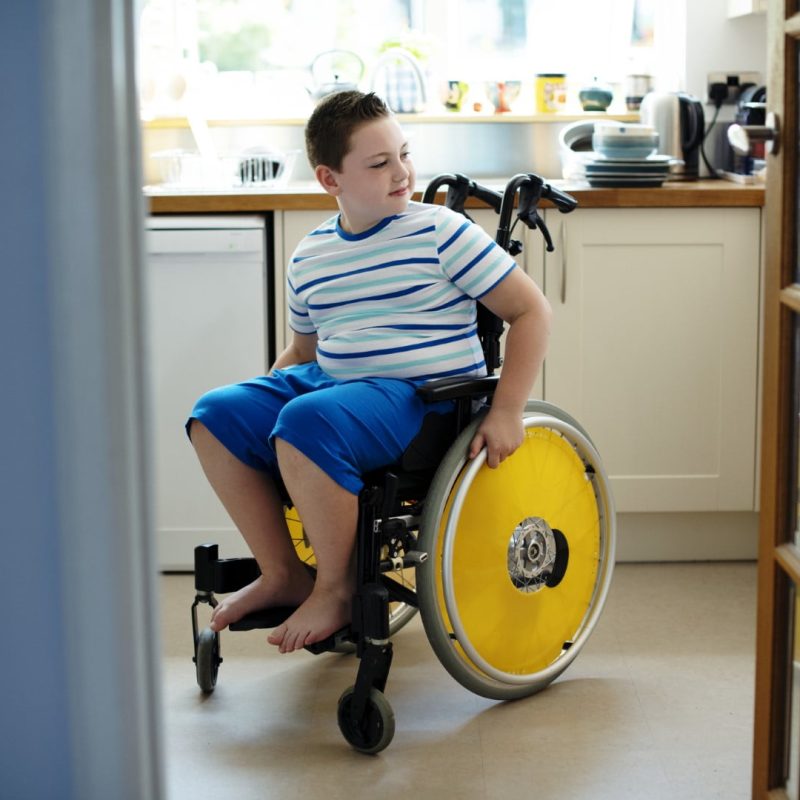 A boy in a wheelchair with yellow wheels sits in a kitchen, looking to his side with a neutral expression. He is wearing a striped shirt and blue shorts.