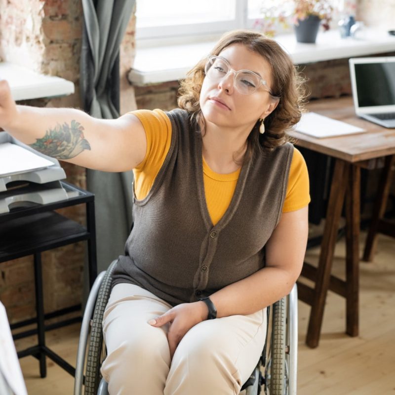 A woman in a wheelchair points at a whiteboard in an office setting, with a laptop and documents on a desk in the background.
